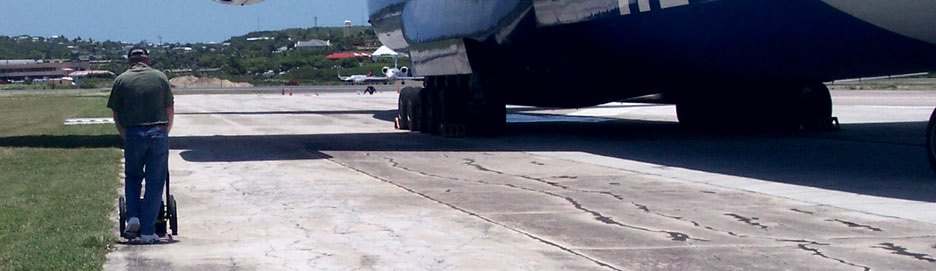 A surveyor pushing a ground-penetrating radar cart along an airport runway beside a parked cargo aircraft in Antigua, surveying for voids and sinkholes.