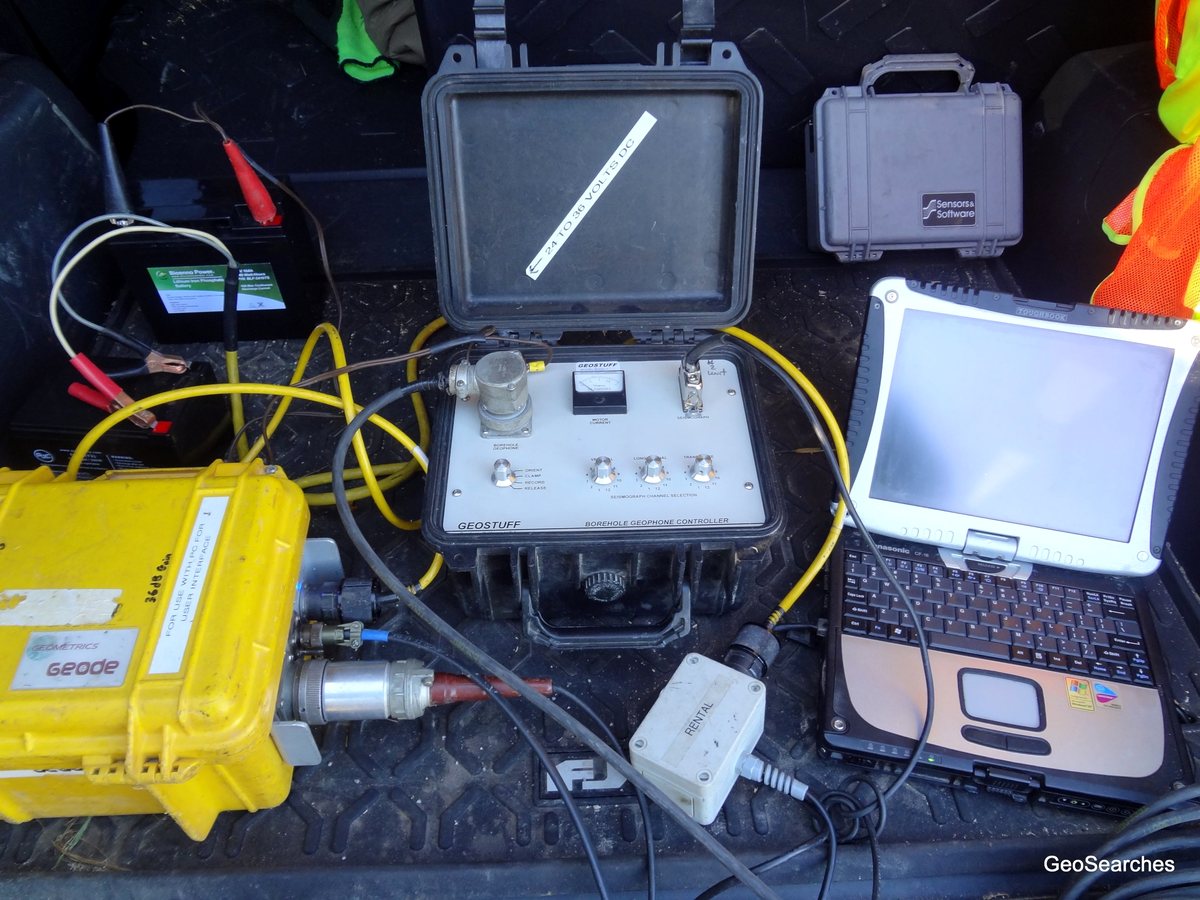 Downhole seismic recording equipment on a vehicle tailgate: a borehole geophone controller with clamp and release switches and a motor current meter, wired to a seismograph and a field laptop.