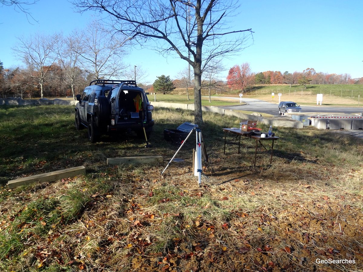 A downhole seismic setup on open ground: a tripod standing over a PVC-cased borehole with the survey vehicle and recording equipment behind it, and a timber shear-wave source beam laid on the ground alongside.