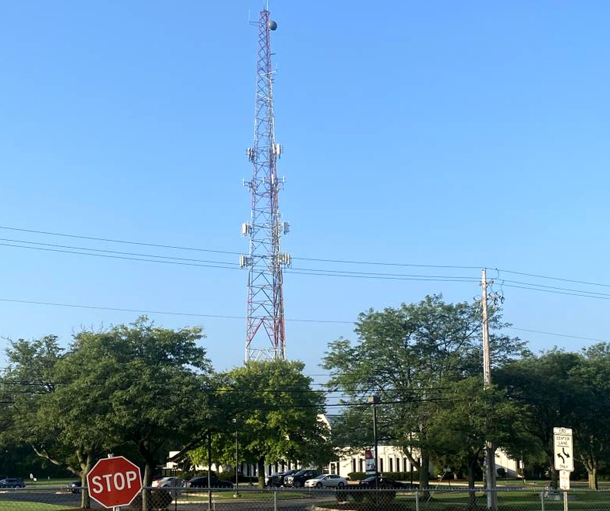 A communications tower carrying multiple antenna arrays rising above a treeline, with overhead electrical distribution lines crossing the foreground above a road.