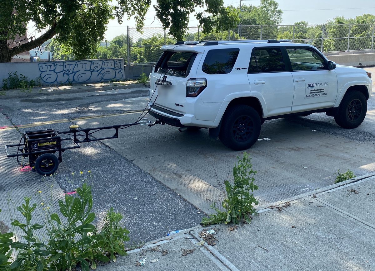 A survey vehicle towing a ground-penetrating radar cart on a tow bar across a concrete deck, collecting a continuous line at driving pace.