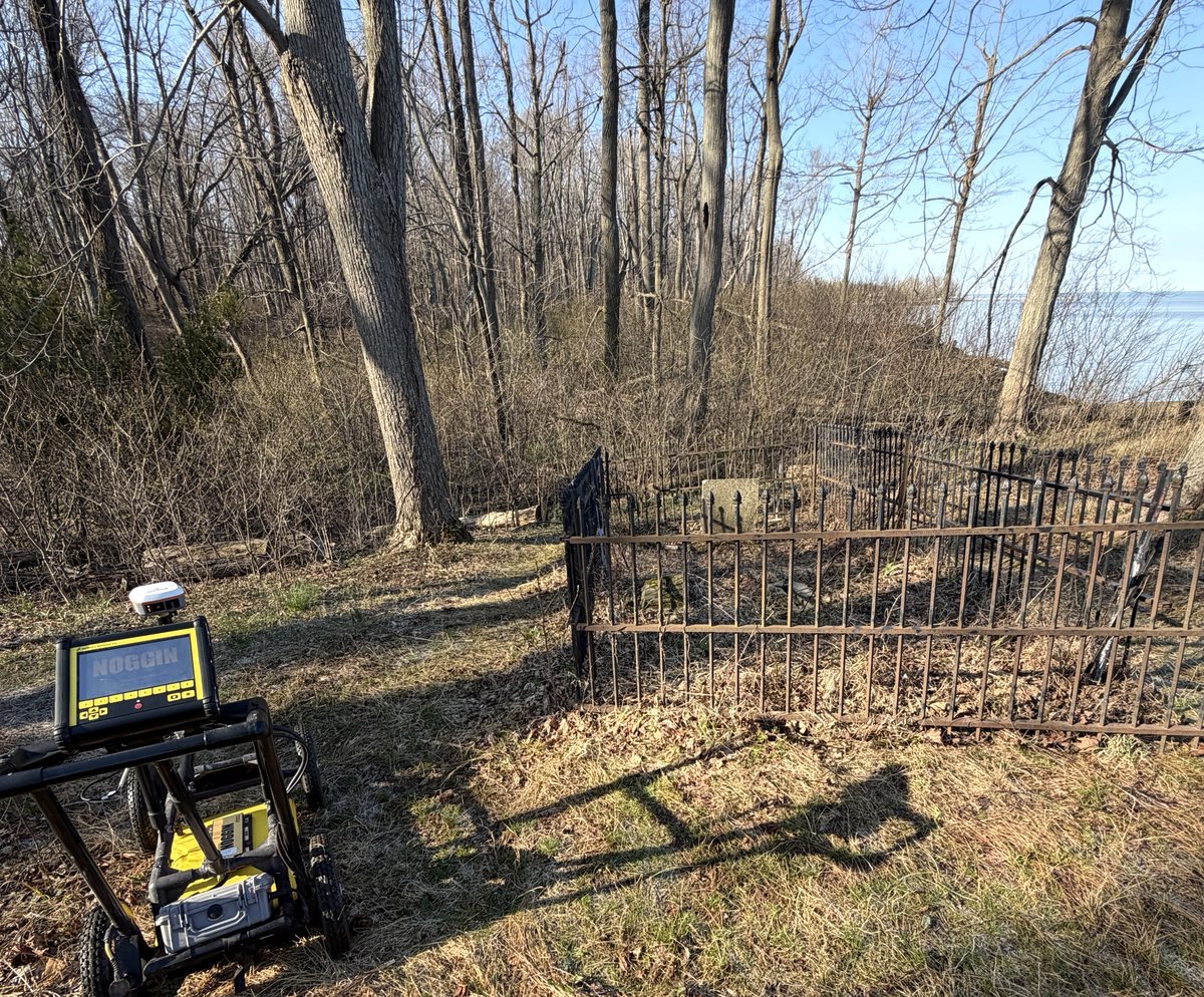A ground-penetrating radar cart standing beside a small wrought-iron-fenced homestead burial plot on a wooded bluff, with Lake Erie visible beyond the trees.