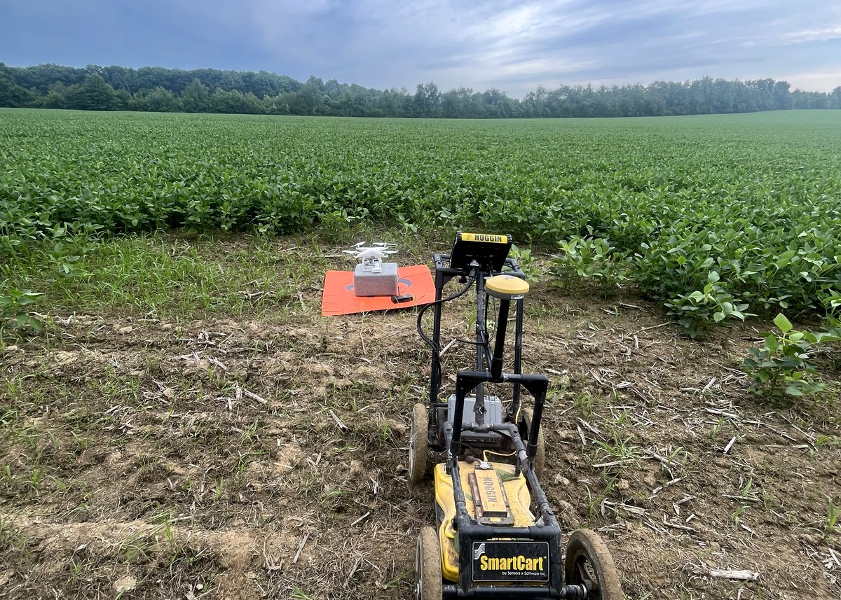 A Noggin SmartCart ground-penetrating radar system with an RTK GNSS antenna standing on the headland of a soybean field, a survey drone on its landing pad behind it.