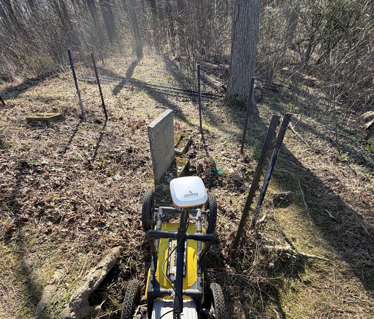 A ground-penetrating radar cart with a GNSS antenna being pushed between weathered headstones inside an iron-fenced burial plot in woodland.