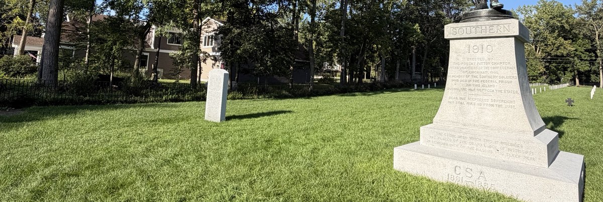 The Confederate Stockade Cemetery on Johnson's Island, Ohio: the 1910 bronze monument on its granite pedestal, an inscribed stone, and rows of white grave markers behind the iron fence.