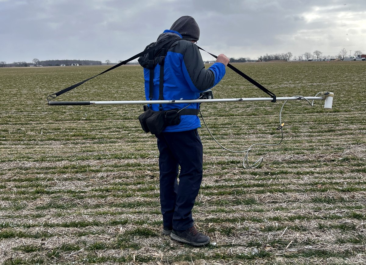 A GeoSearches surveyor walking a stubble field in winter carrying a cesium vapour magnetometer on a horizontal boom, searching for abandoned well casings.