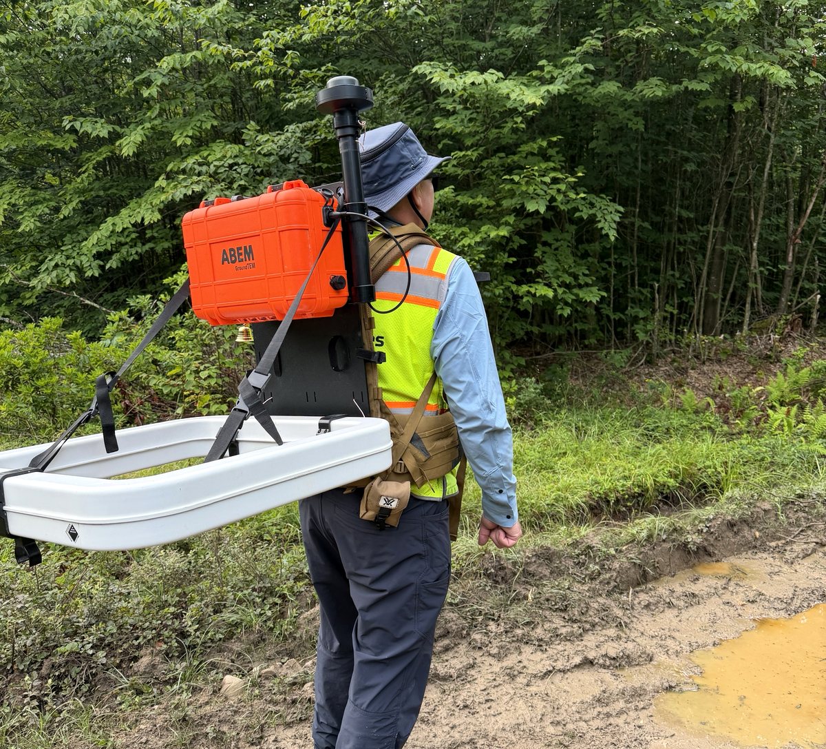 A GeoSearches surveyor carrying a backpack-mounted ABEM GroundTEM time-domain electromagnetic system along a wet forest track, with the transmitter loop on a shoulder frame and an RTK GNSS antenna on a mast above it.