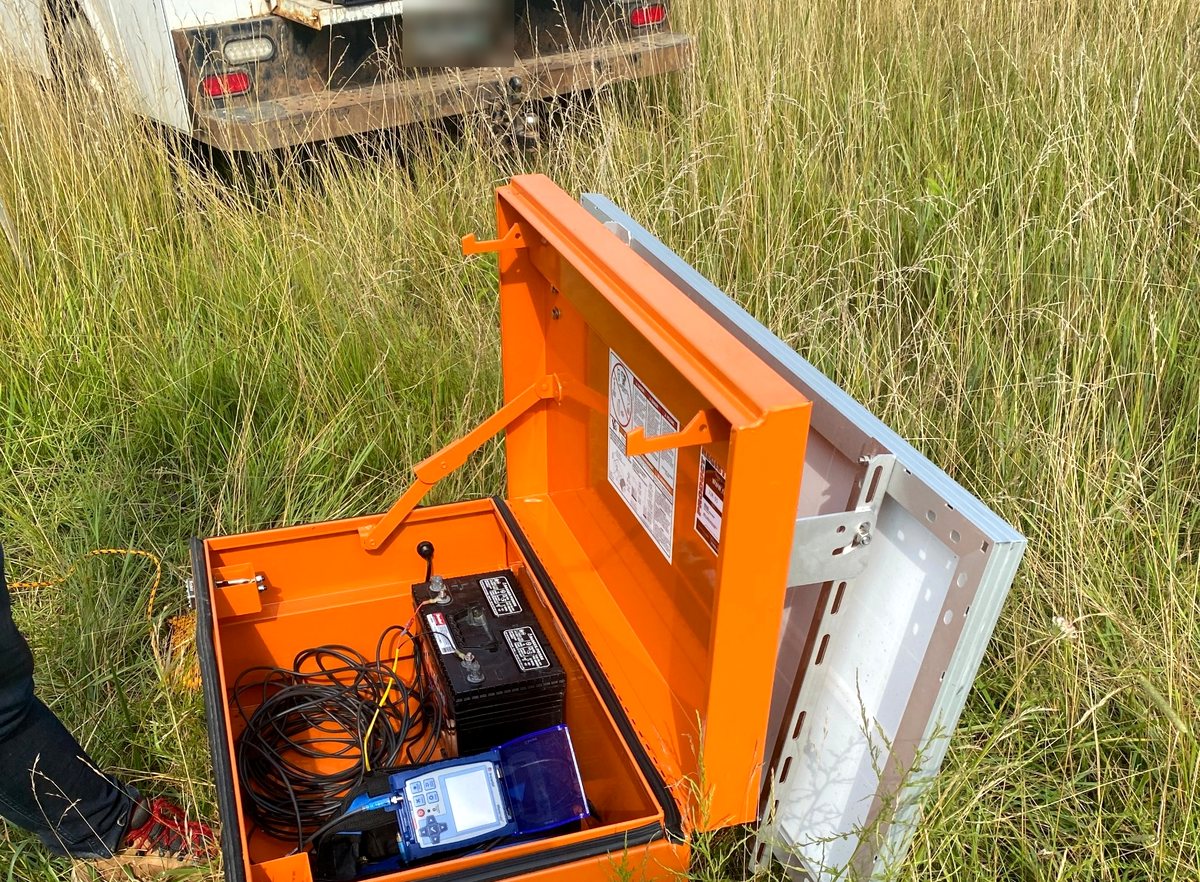 An open weatherproof case on the ground containing a seismograph, a deep-cycle battery and coiled geophone cable, with the solar panel mounted on the lid.
