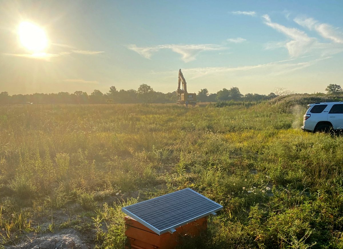 A solar-powered vibration monitoring station in a weatherproof case standing in rough grass, with an excavator working on the horizon beyond it in low morning sun.